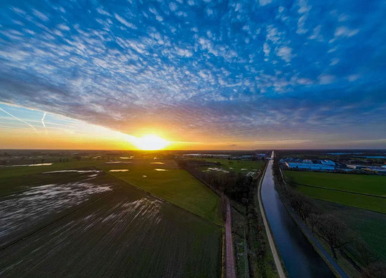 Aerial view of a sunset over a rural landscape with fields, a canal, and scattered clouds captured by the DJI Mini 3 Pro.