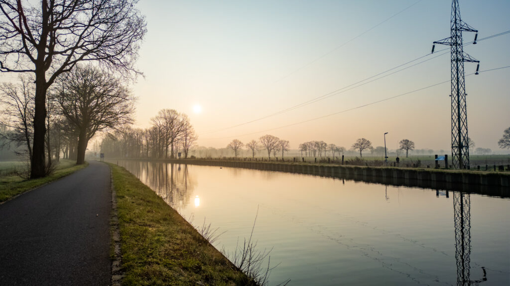 Sunrise over a tranquil canal next to a country road with power lines and trees.