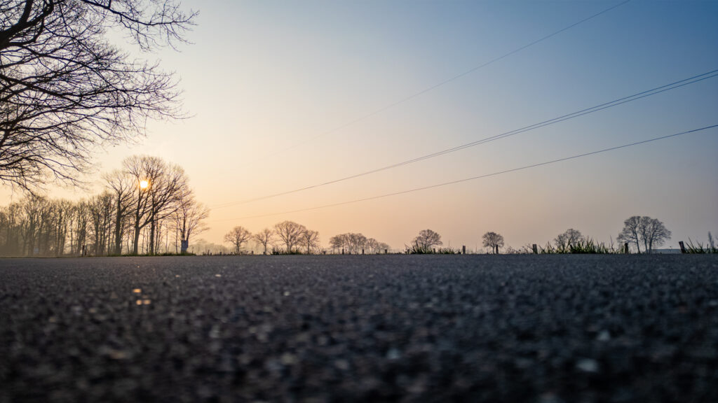 Sunrise with hazy sky seen from a low angle on an empty road surrounded by trees.