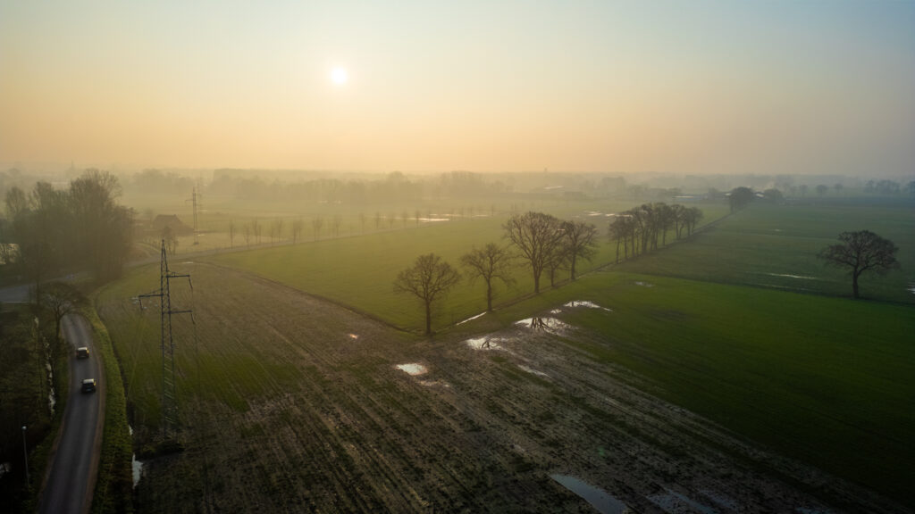 Hazy sunrise over a tranquil countryside with a road winding beside fields and scattered trees.