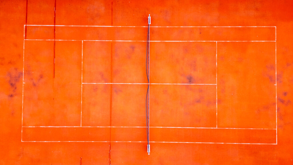 Drone's view of an empty, red tennis court with white lines marking the boundaries and sections of the playing area.