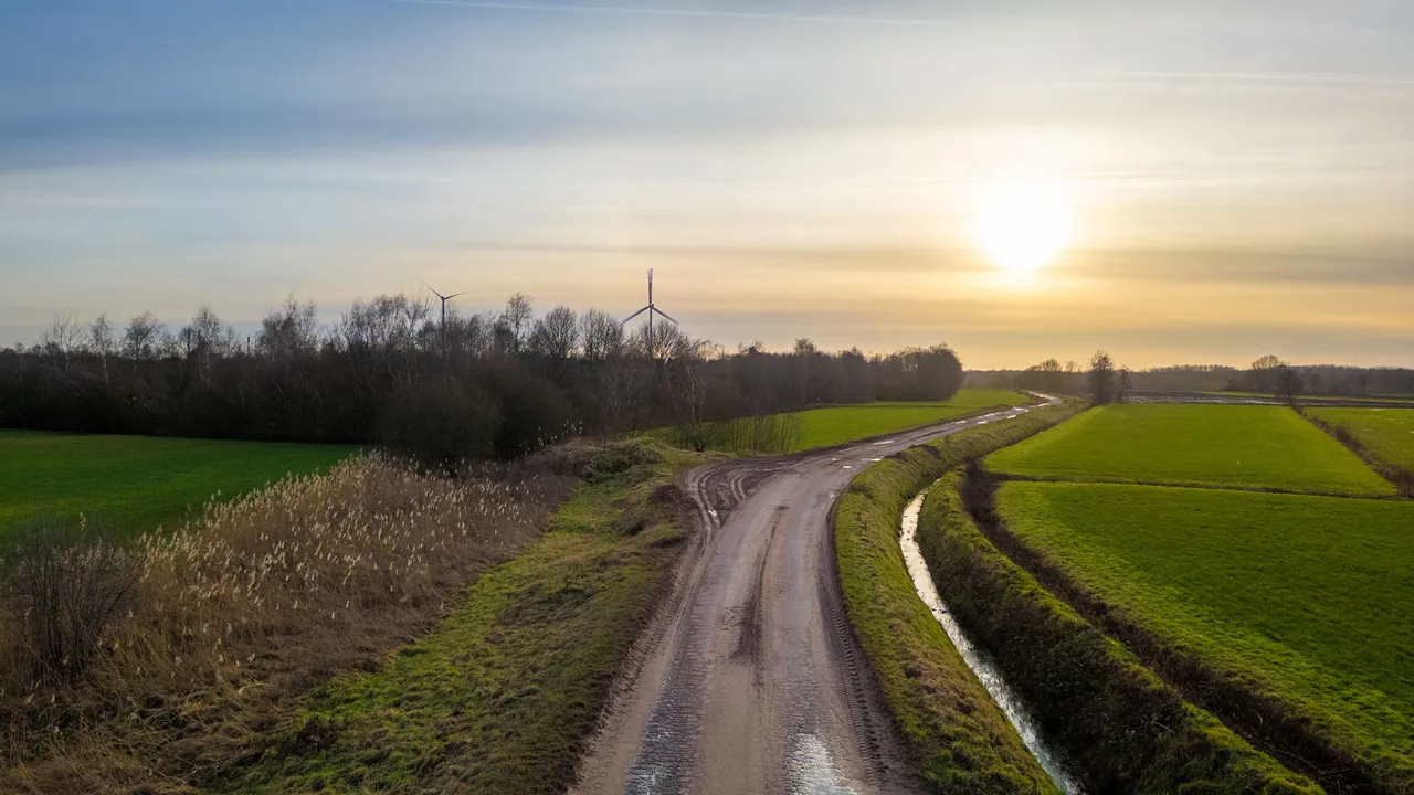 A meandering country road at sunset with a wind turbine in the background, captured through the unfiltered lens of a DJI Mini 3 Pro.