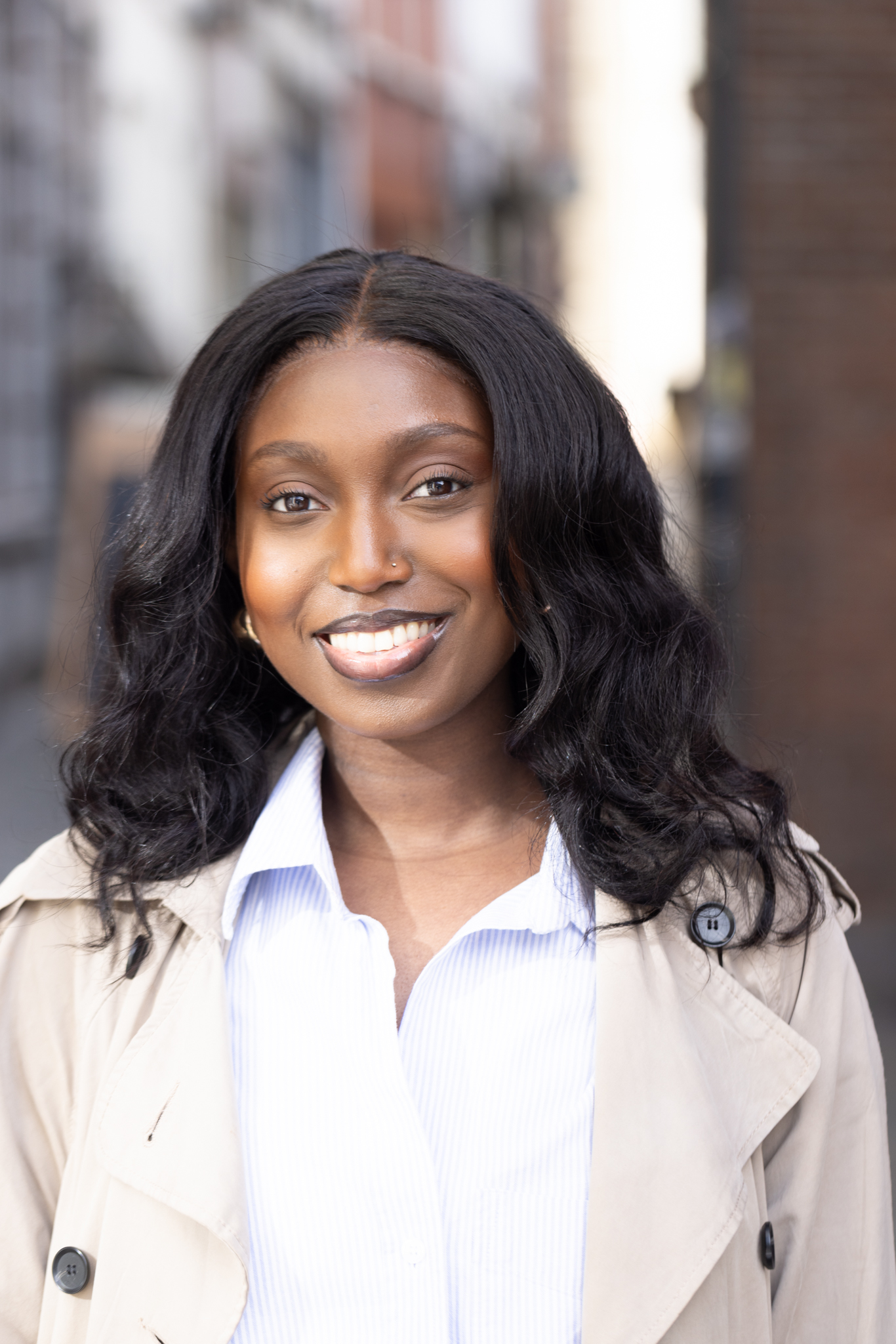 A smiling woman wearing a beige trench coat and a blue shirt, captured with a Canon R5 outdoors during her lunch break adventure.